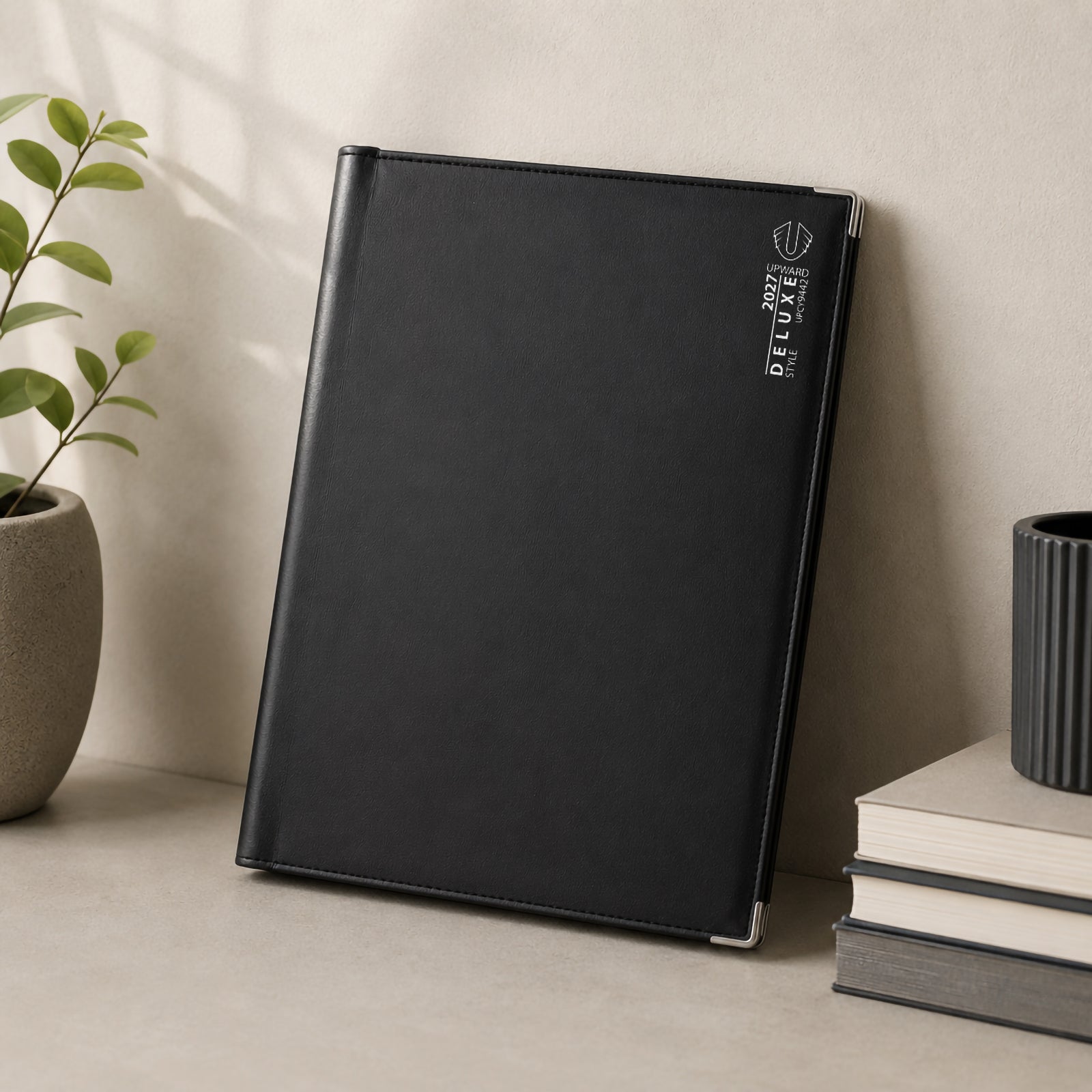 The Upward Deluxe 2027 A4 Week To View Diary stands upright beside stacked books and decorative greenery. Its elegant black PU leather cover, clean stitching and protective silver corner details present a refined diary designed for everyday desk planning throughout 2027.