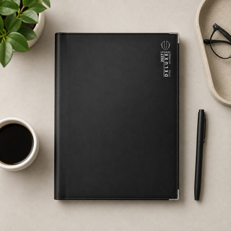 Overhead view of the Upward Deluxe 2027 A4 Week To View Diary resting on a minimalist workspace beside a coffee cup, pen, glasses and indoor plant. The spacious A4 format and premium black cover create an organised, professional planning solution for offices and home workspaces.