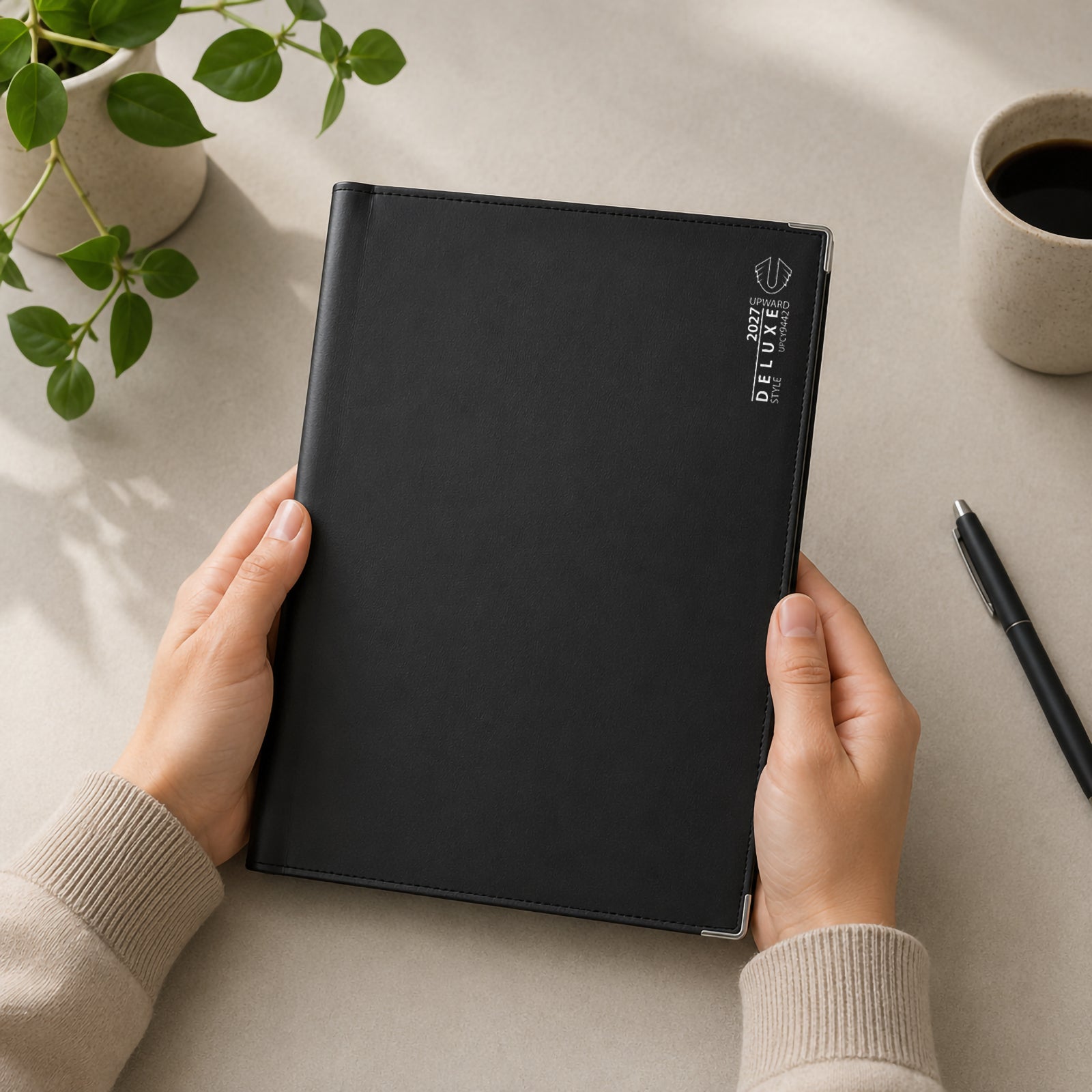 A person holds the Upward Deluxe 2027 A4 Week To View Diary featuring a premium black PU leather cover with silver branding and reinforced metal corner protectors. Positioned on a modern desk with a coffee cup and pen, the diary showcases its generous A4 size for professional weekly planning.