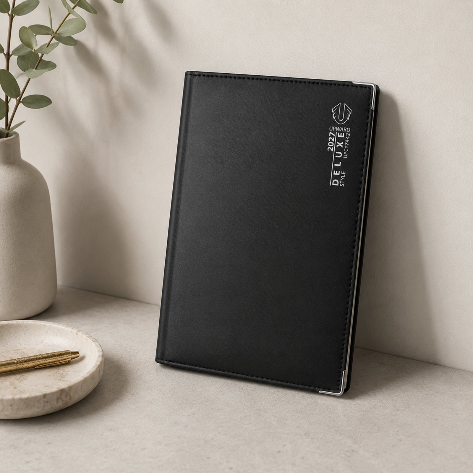 The Upward Deluxe 2027 A5 Week To View Diary stands upright on a stylish office shelf alongside decorative plants and workspace accessories. Its sleek black PU leather finish and silver detailing present a sophisticated planner suited to professional and personal organisation.