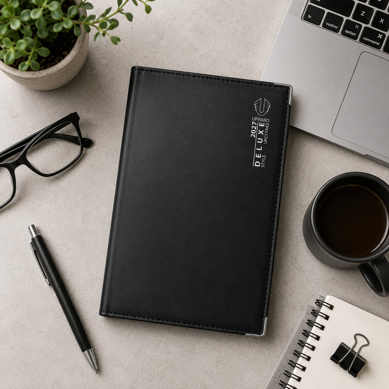Top down view of the Upward Deluxe 2027 A5 Week To View Diary placed on a modern desk with a laptop, glasses, coffee cup, notebook and stationery. The premium black cover and reinforced metal corner protectors highlight its durable executive design.