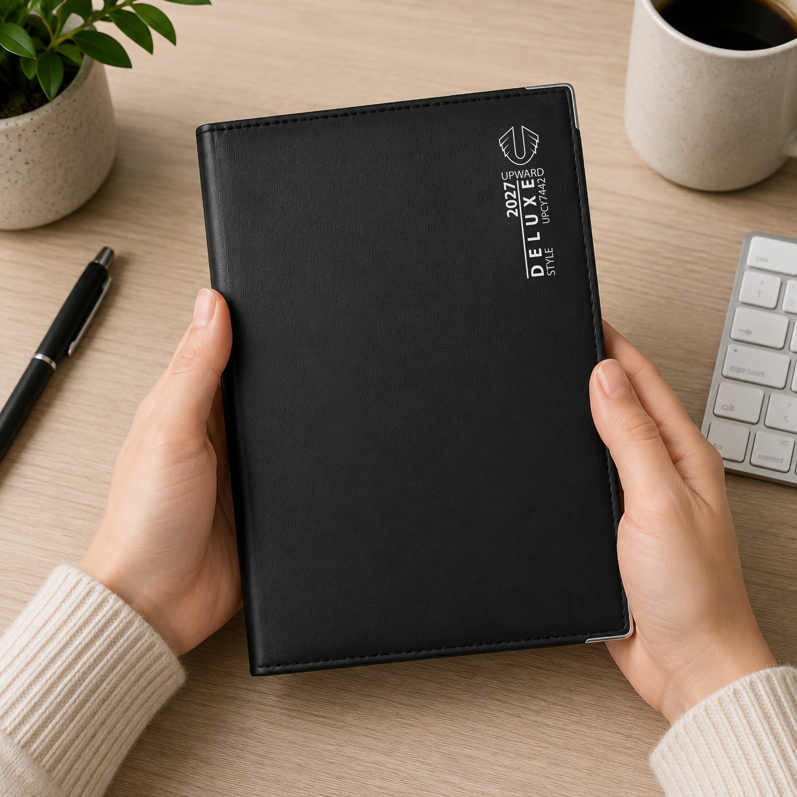 A person holds the Upward Deluxe 2027 A5 Week To View Diary featuring a premium black PU leather cover with elegant silver branding. Positioned in a contemporary workspace beside a keyboard, coffee mug and pen, the diary demonstrates its portable size and professional appearance for everyday planning.