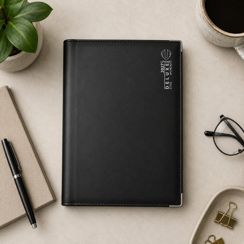 Overhead desktop view of the Upward Deluxe 2027 B5 Day To Page Diary positioned beside a notebook, pen, coffee cup and office accessories. The premium black cover and reinforced metal corner protectors showcase a stylish diary designed for organised daily scheduling.