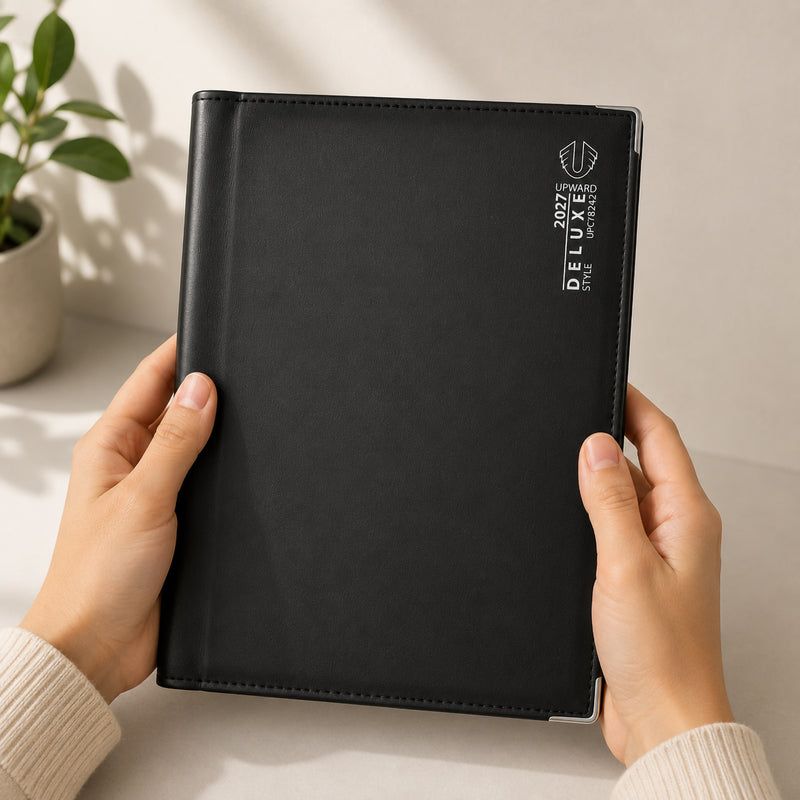 A person holds the Upward Deluxe 2027 B5 Day To Page Diary featuring a smooth black premium PU leather cover with elegant silver branding. The diary is presented in a modern workspace, highlighting its comfortable size, refined appearance and professional finish for everyday planning.