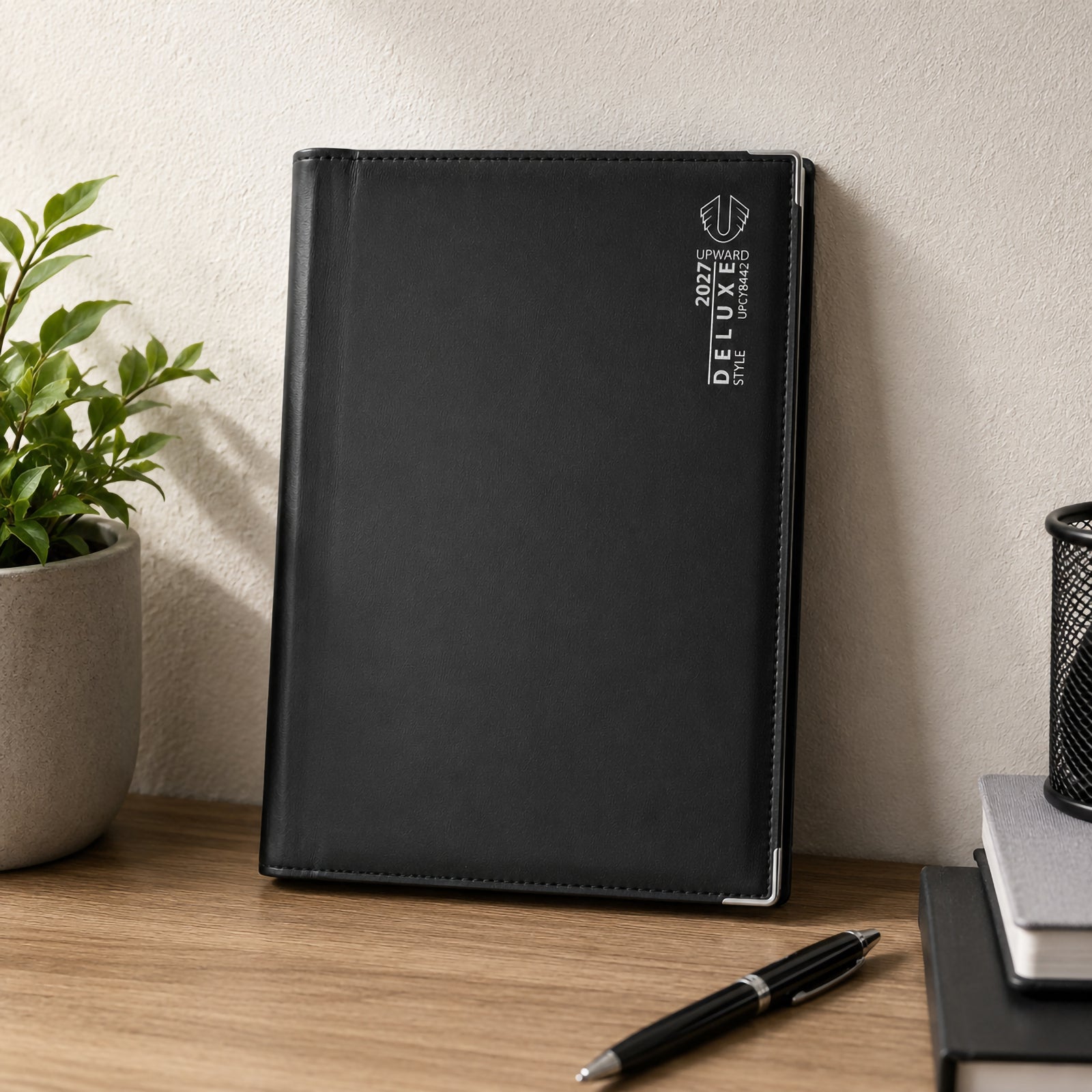 The Upward Deluxe 2027 B5 Diary stands upright on a tidy desk beside stationery and indoor plants. The elegant black PU cover, refined silver branding and reinforced corner protectors create a professional look suited to daily planning throughout the year.