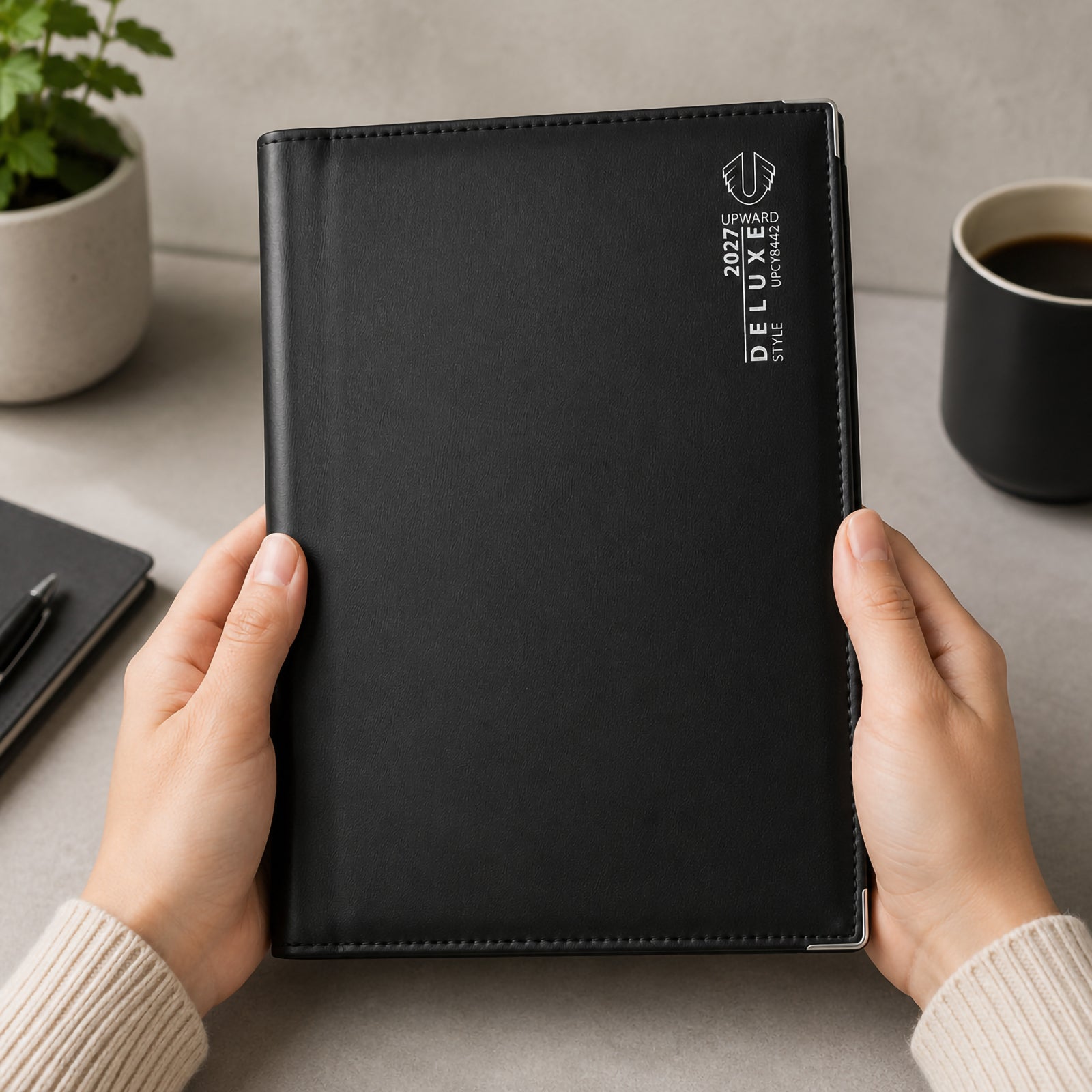 A person holds the Upward Deluxe 2027 B5 Week To View Diary featuring a smooth black premium PU leather cover with silver branding. The diary is shown in a modern workspace with coffee, stationery and indoor plants, highlighting its comfortable size, professional appearance and quality finish.