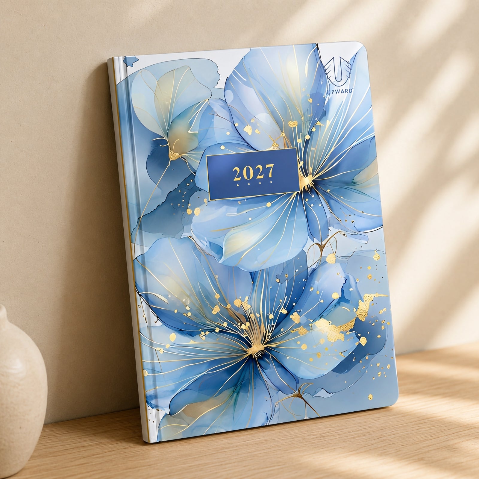 The Upward Fashion 2027 A5 Hardcover Day To Page Diary stands upright on a timber shelf with soft natural lighting, showcasing the luxurious Water Colour floral cover, rounded corners and premium hardcover construction designed for stylish year round planning.