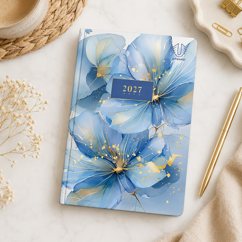 Flat lay view of the Upward Fashion 2027 A5 Hardcover Day To Page Diary displayed beside a gold pen, coffee cup and decorative accessories, highlighting the sophisticated blue floral Water Colour artwork and compact A5 hardcover design.