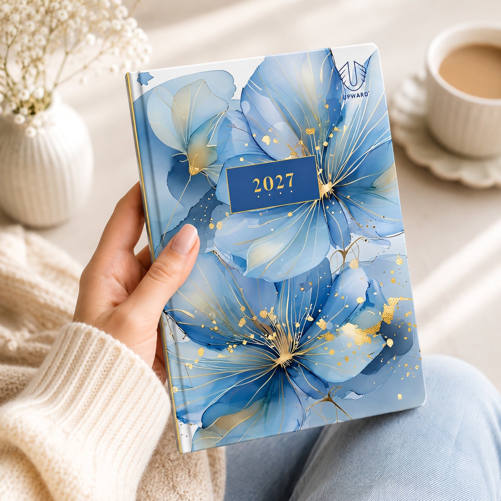 A person holds the Upward Fashion 2027 A5 Hardcover Day To Page Diary featuring an elegant Water Colour floral design with layered blue flowers, delicate gold detailing and a premium hardcover finish, creating a stylish planner suitable for everyday personal and professional organisation.