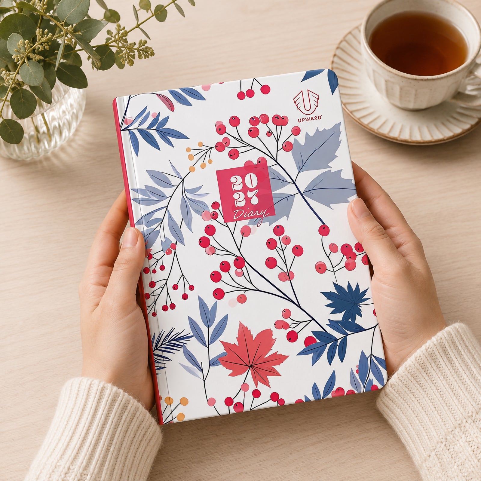 A person holds the Upward Fashion 2027 A5 Hardcover Week To Opening Diary featuring an elegant Begonia inspired botanical cover with vibrant red berries, soft blue foliage and a premium hardcover finish, highlighting its stylish design for everyday planning at home or work.
