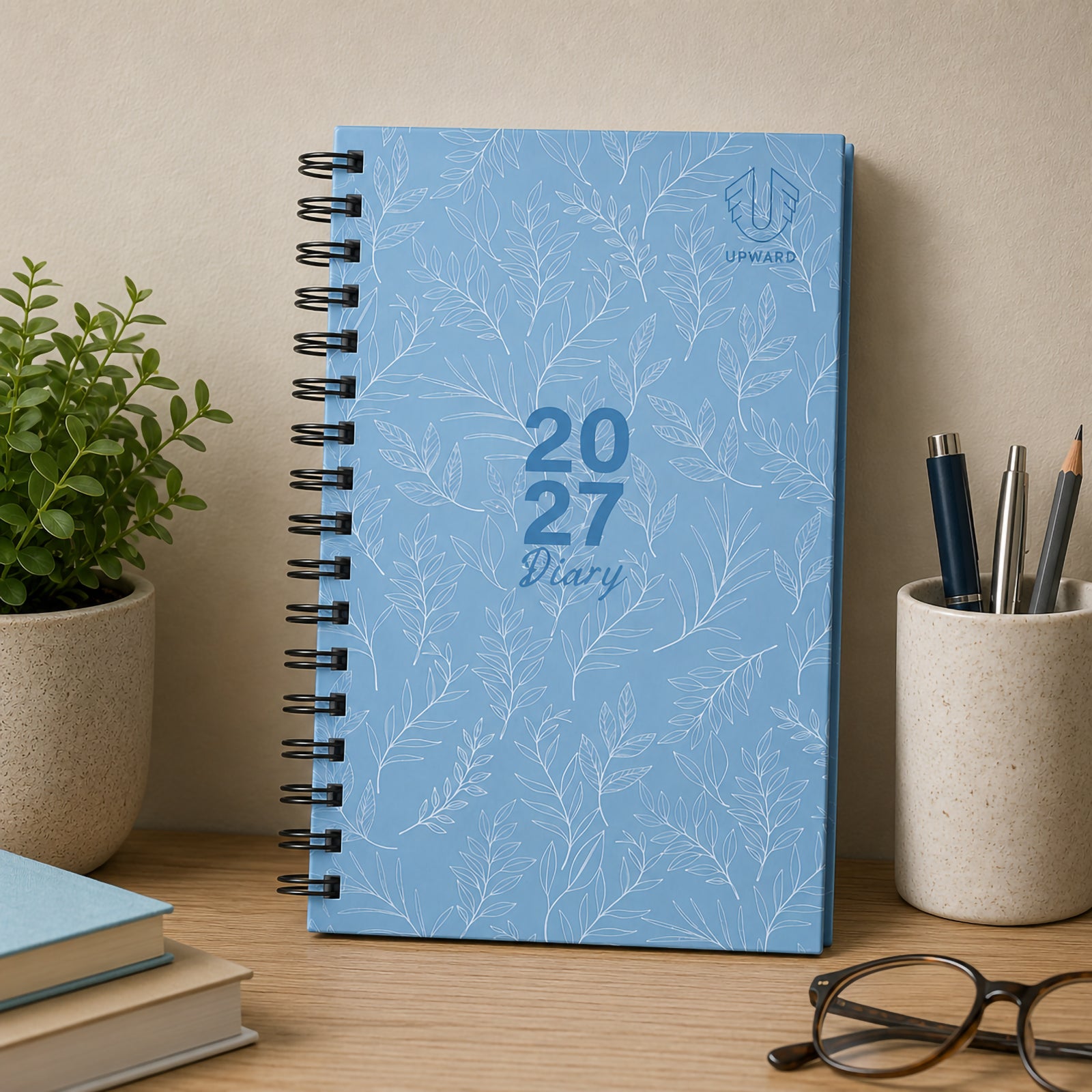 Front cover lifestyle image featuring the Upward Fashion 2027 Blue Diary displayed beside books, pens and office accessories. The soft blue botanical design and durable hardcover create a professional appearance suitable for business, study or personal organisation.