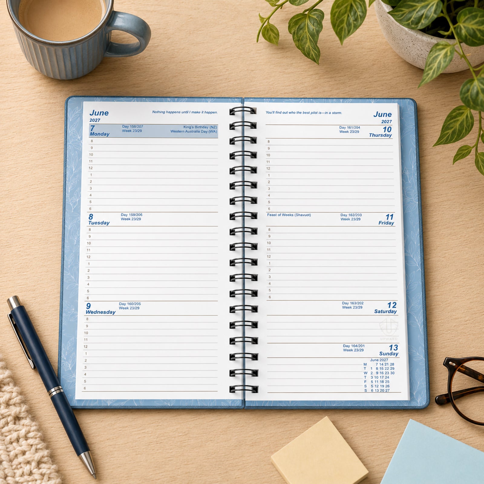 The Upward Fashion 2027 A6/7 Blue Diary is displayed open on a timber desk, showcasing its week to view layout with generous writing space for appointments, meetings and daily tasks. Surrounded by stationery, a coffee cup and indoor plants, the diary presents an organised and productive workspace.