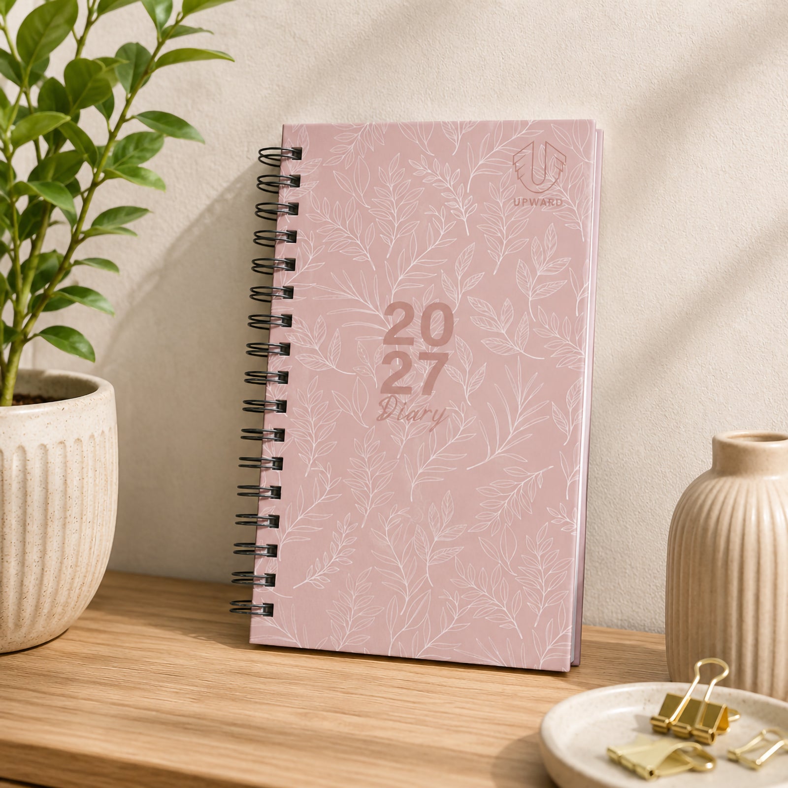 Front cover displayed on a neatly styled desk with decorative plants and office accessories. The pastel pink hardcover with delicate botanical illustrations creates a refined and calming appearance suitable for everyday planning.