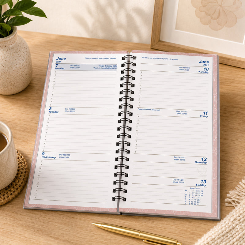 Lifestyle image showing the Upward Fashion 2027 A6/7 Pink Diary opened on a timber desk. The spacious week to view pages provide generous writing space for appointments, meetings and reminders, with a coffee cup and stationery creating a warm and organised workspace.