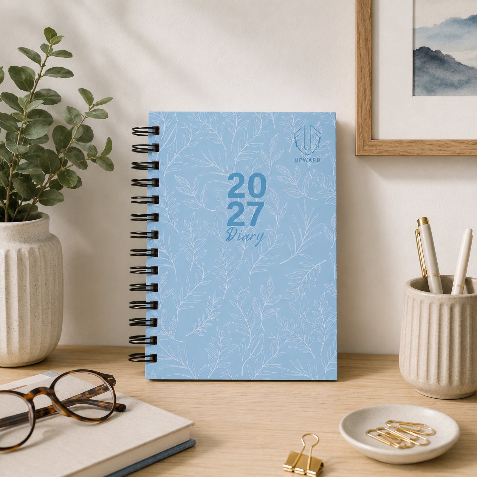 The diary displayed upright on a tidy desk surrounded by stationery and decorative plants. The elegant blue hardcover design and wire O binding create a practical yet stylish planning solution for home or office environments.