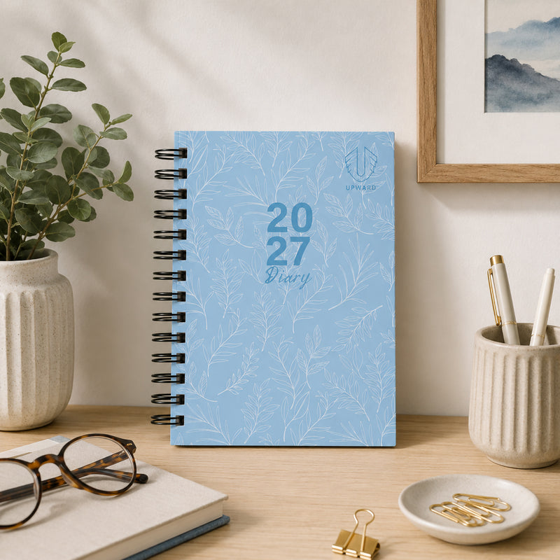 The diary displayed upright on a tidy desk surrounded by stationery and decorative plants. The elegant blue hardcover design and wire O binding create a practical yet stylish planning solution for home or office environments.