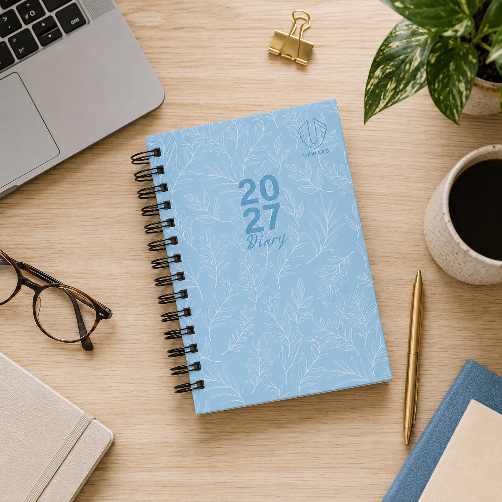 Overhead workspace scene featuring the closed blue diary positioned beside a laptop, glasses, notebook, coffee cup and desk accessories. The soft blue cover with delicate botanical pattern presents a professional and calming appearance for everyday planning.