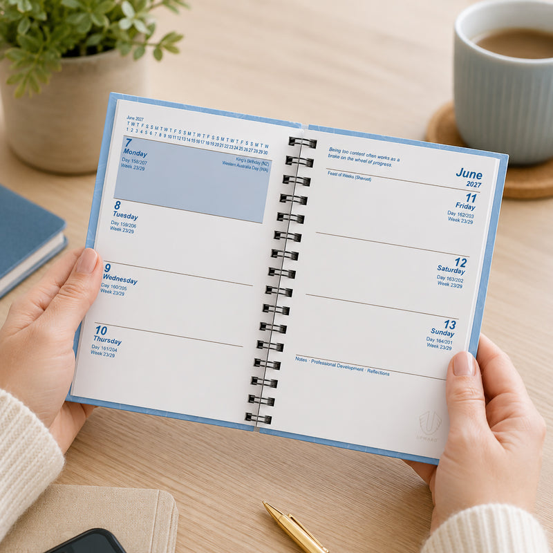 Lifestyle image showing the Upward Fashion 2027 A7 Blue Diary opened to the weekly planning pages. The diary is held in hand on a light timber desk, highlighting the spacious week to view layout with dedicated sections for appointments, notes and important dates.