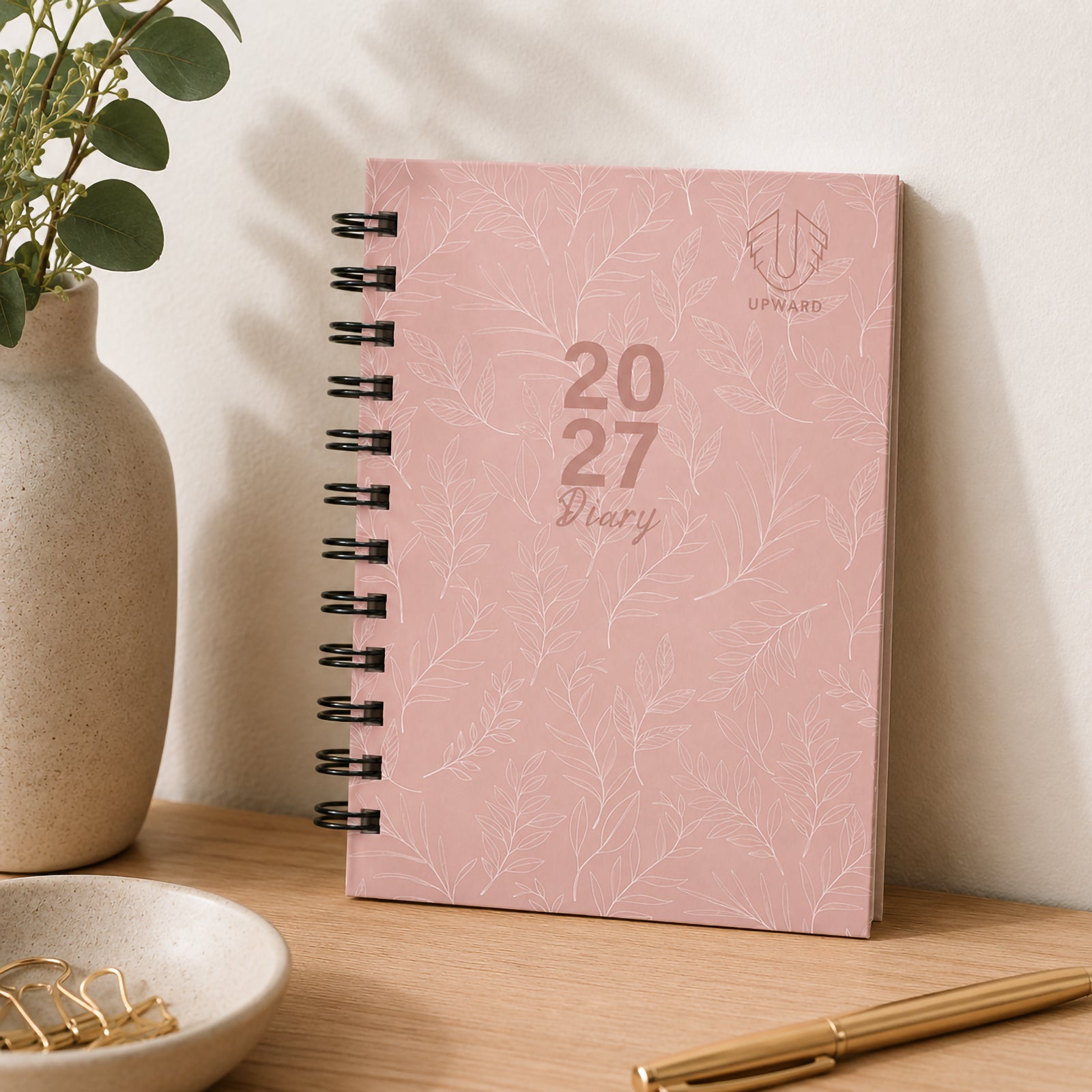 The diary standing upright on a desk against a neutral background with decorative greenery and premium stationery. The blush pink hardcover and subtle leaf pattern provide a stylish yet professional look for everyday organisation.