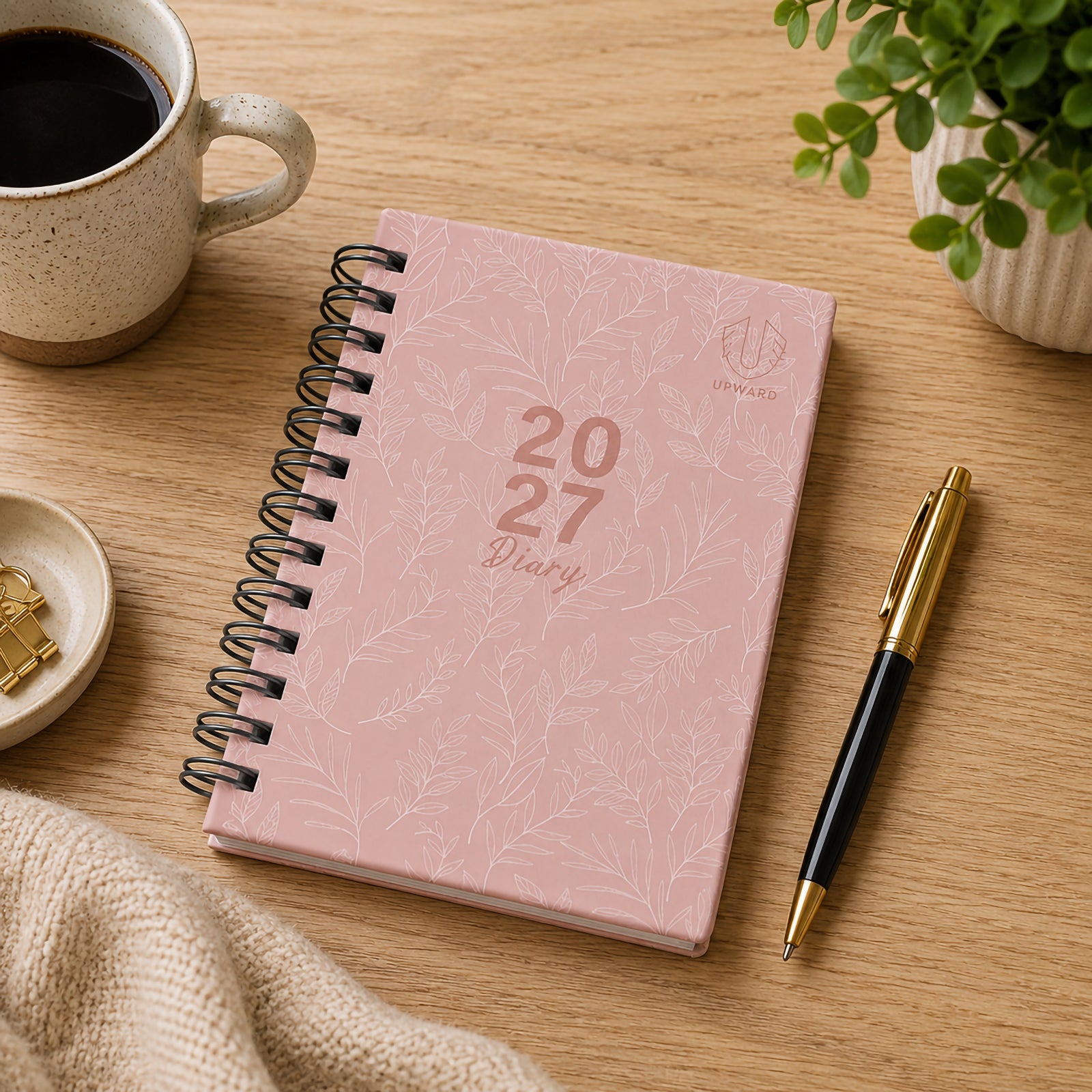 Overhead lifestyle image of the closed pink diary resting on a wooden desk beside a coffee cup, gold and black pen and indoor plant. The elegant botanical cover pattern creates a calm and sophisticated appearance suitable for professional and personal planning.