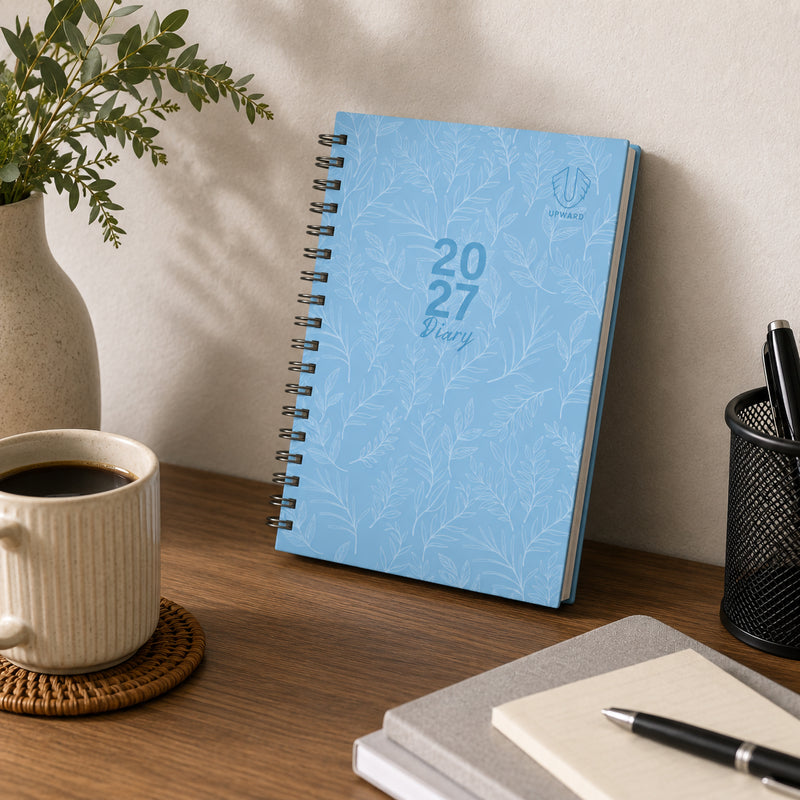 The 2027 Blue Sky diary displayed upright on a desk near office stationery and a coffee mug. The stylish hardcover and Wire O binding showcase a balance of functionality and modern design for everyday use.
