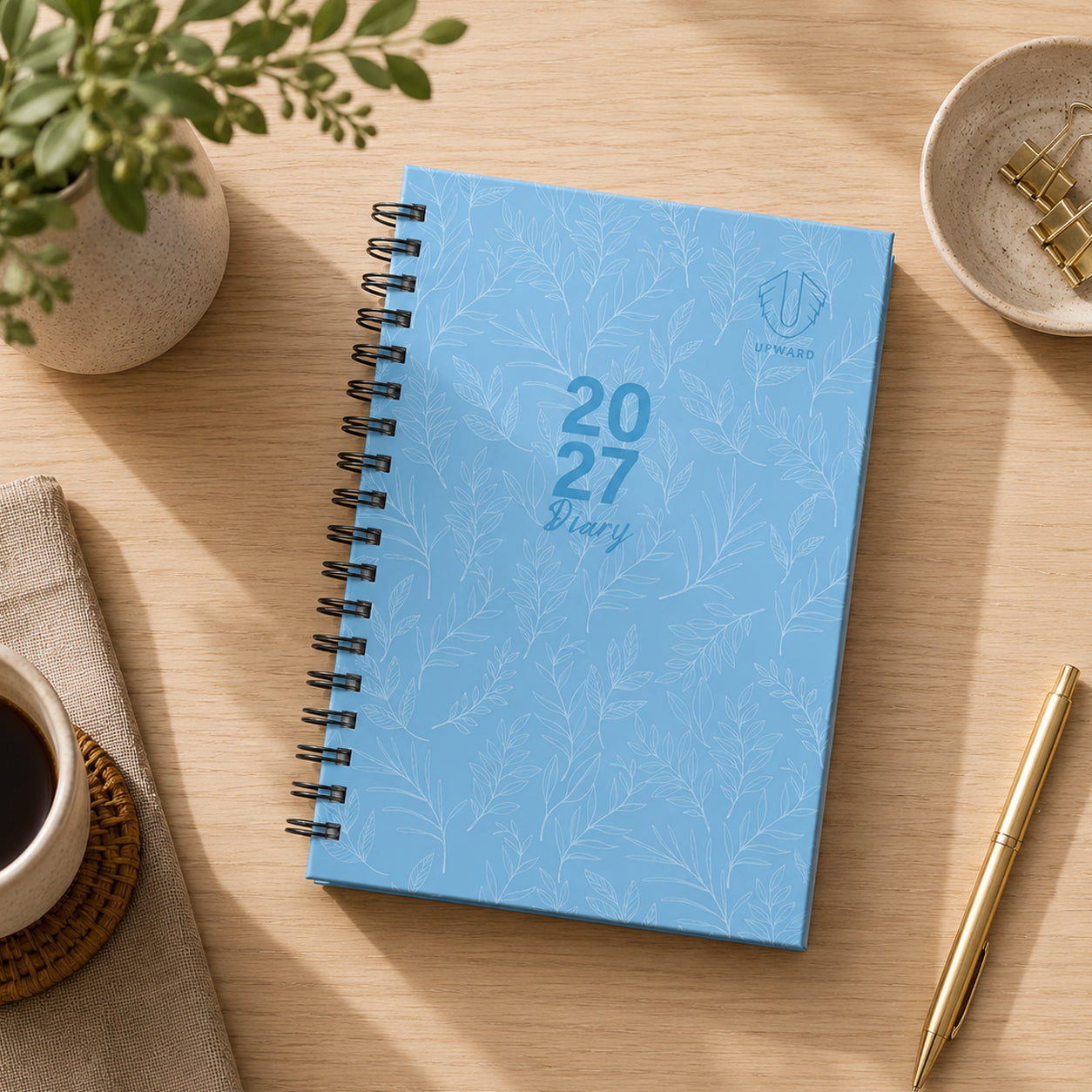 Top down lifestyle view of the blue hardcover diary resting on a desk beside a coffee cup, gold pen and decorative greenery. The elegant botanical pattern and soft blue finish create a calming and professional planning companion.