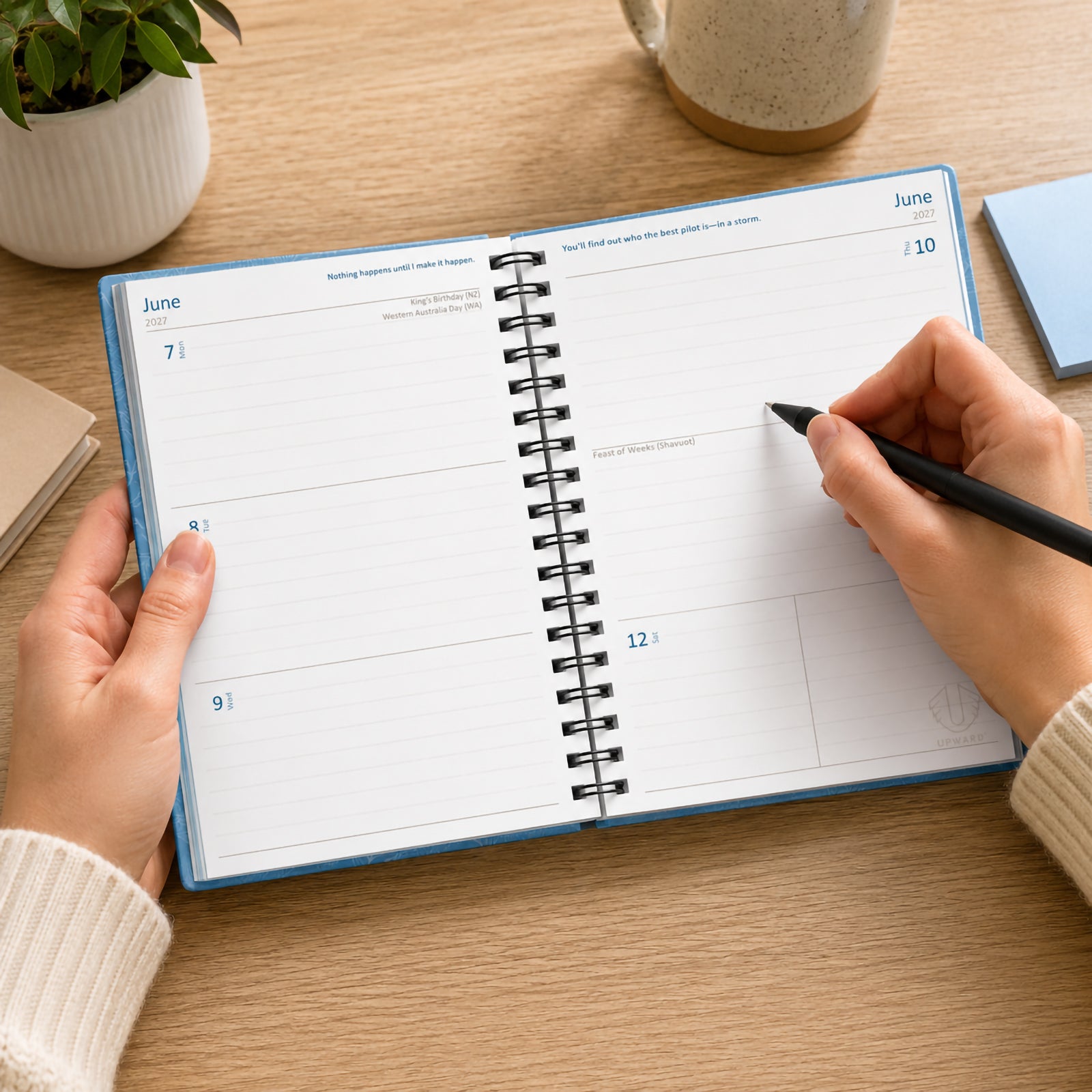 The Upward Fashion 2027 Blue Sky diary opened to a week to view spread on a timber desk. Hands hold the diary while writing notes, demonstrating the spacious weekly layout, flat opening design and practical planning format for work, study or personal organisation.