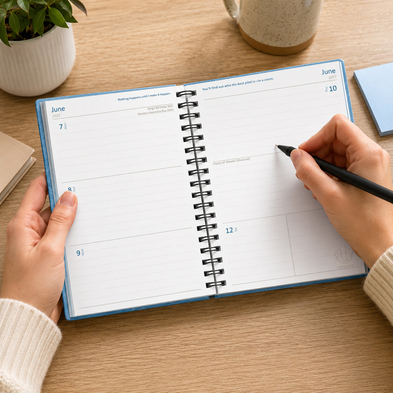 The Upward Fashion 2027 Blue Sky diary opened to a week to view spread on a timber desk. Hands hold the diary while writing notes, demonstrating the spacious weekly layout, flat opening design and practical planning format for work, study or personal organisation.