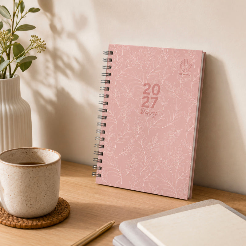 Pink botanical diary standing upright on a desk beside a ceramic coffee cup, decorative vase and notebooks. The elegant hardcover design complements modern home offices and study spaces while offering practical year round planning.