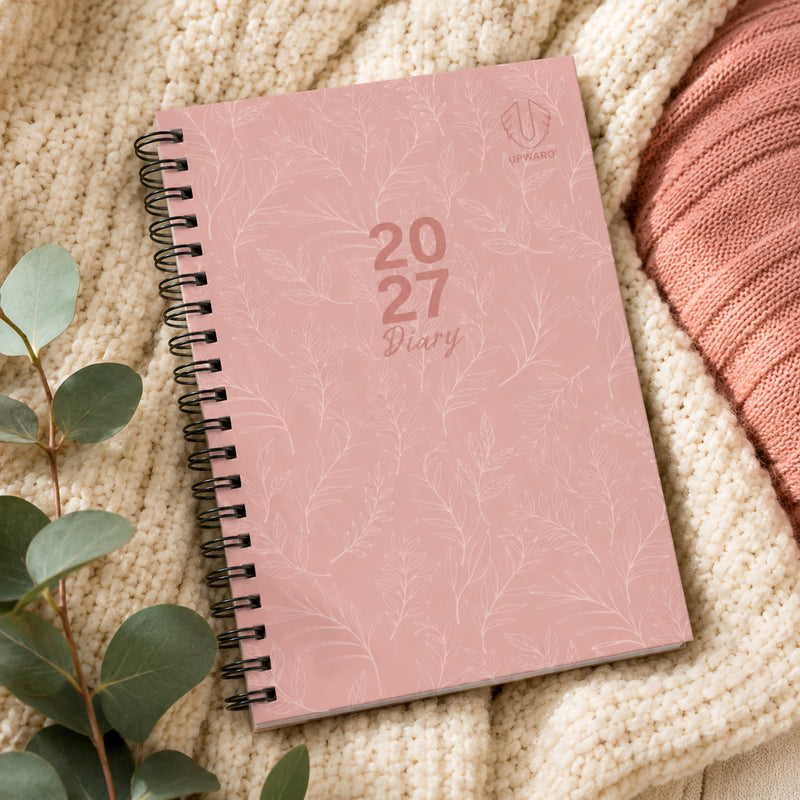 The Upward Fashion 2027 B6 diary displayed on a cosy knitted blanket beside eucalyptus leaves. The soft pink hardcover features a delicate botanical pattern and spiral binding, creating a fashionable planning solution for personal and professional organisation.