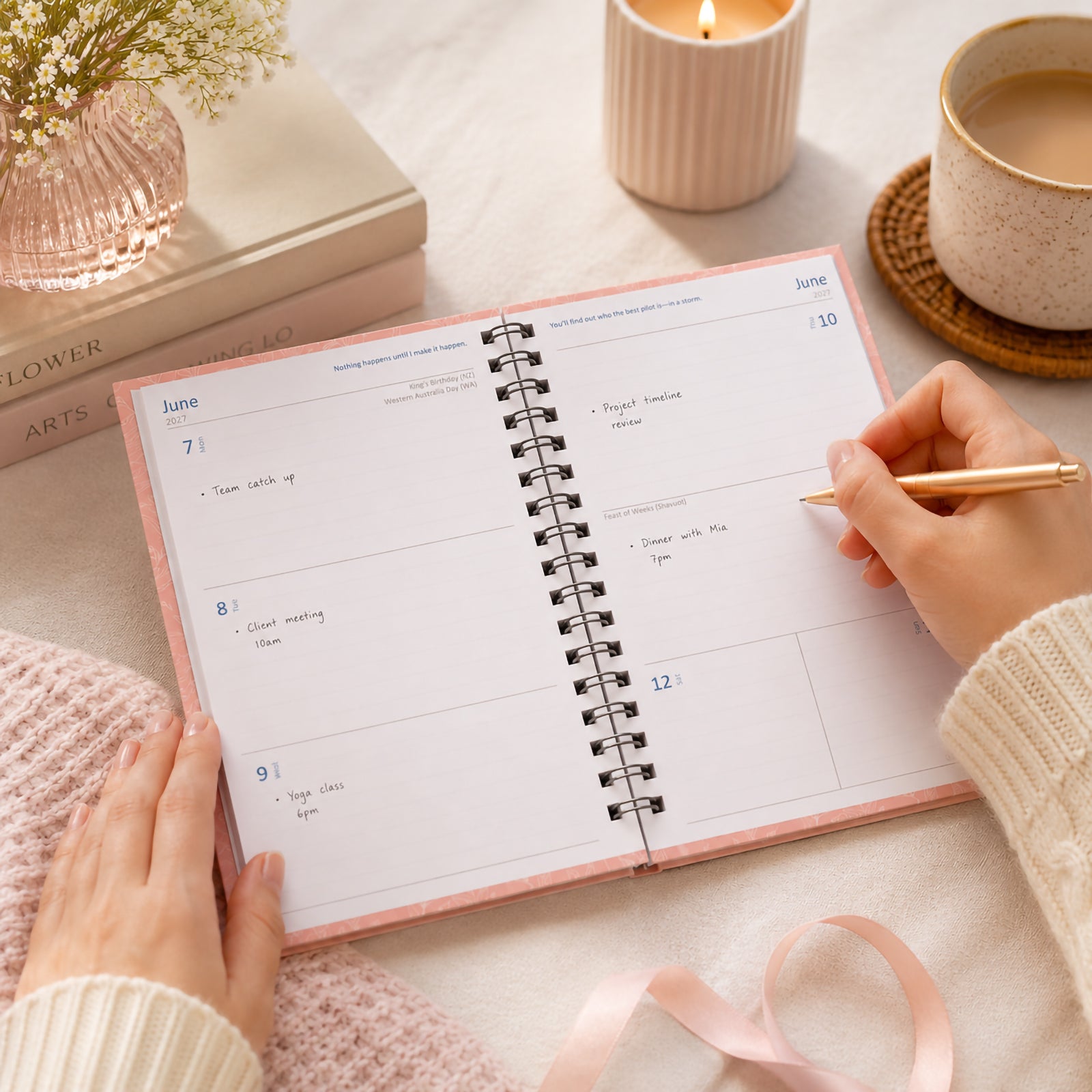 A stylish pink Upward Fashion 2027 diary opened to a weekly planning spread on a soft neutral workspace. A user writes appointments and reminders while surrounded by books, a candle, flowers and a coffee cup, highlighting the practical week to view layout and elegant lifestyle appeal.