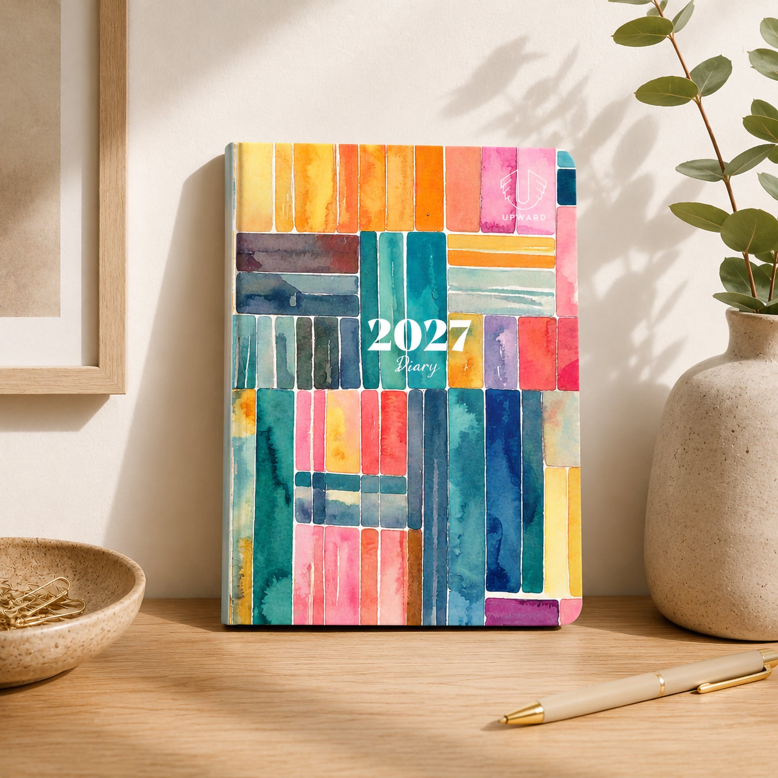 Lifestyle display of the diary standing upright on a desk beside decorative greenery and stationery. The bold artistic cover design brings colour and personality to any workspace while maintaining a professional planning function.