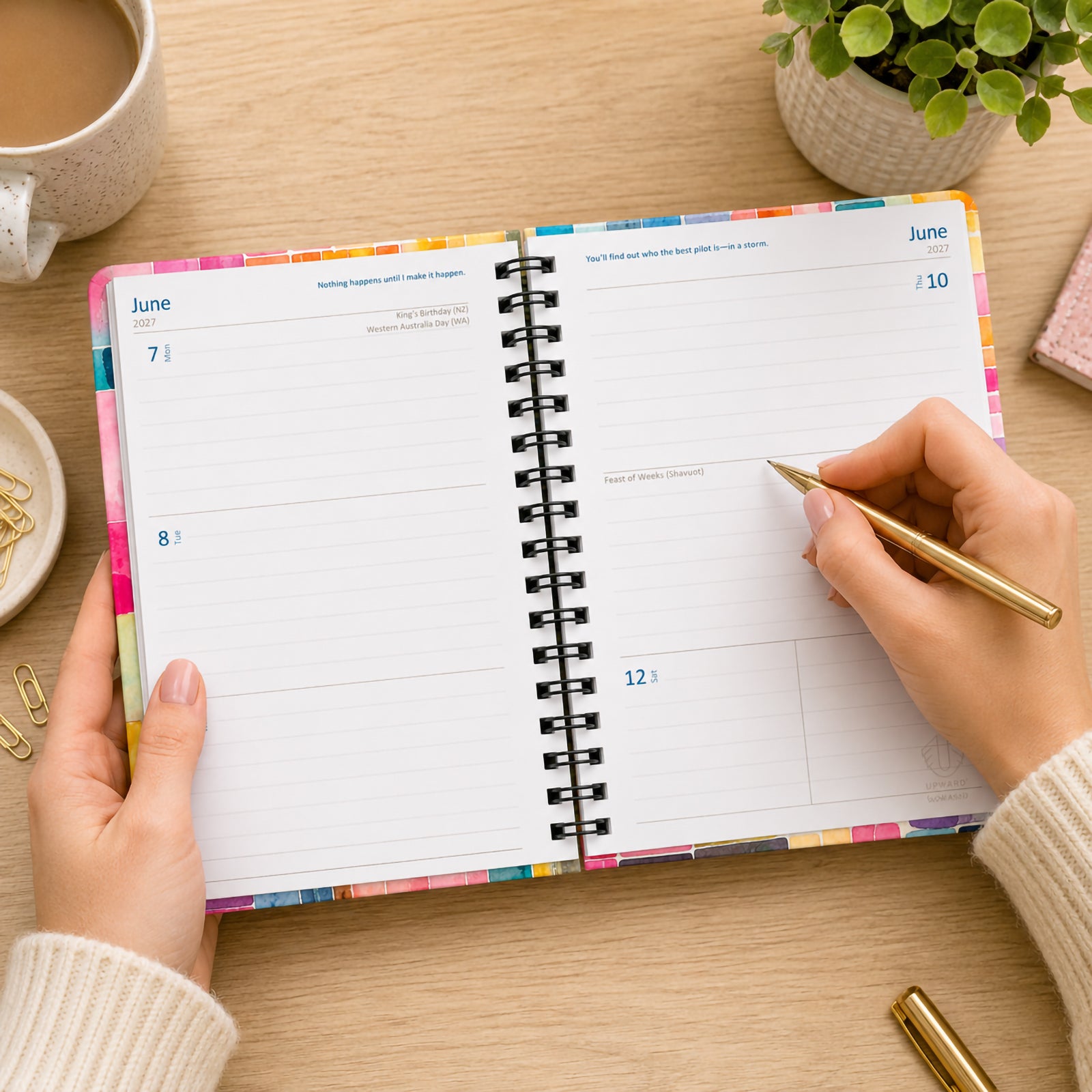 The Upward Fashion 2027 Printed Stripe diary opened on a timber desk while a user writes notes with a gold pen. The week to view layout provides spacious daily sections for appointments, reminders and weekly planning, surrounded by stylish office accessories.