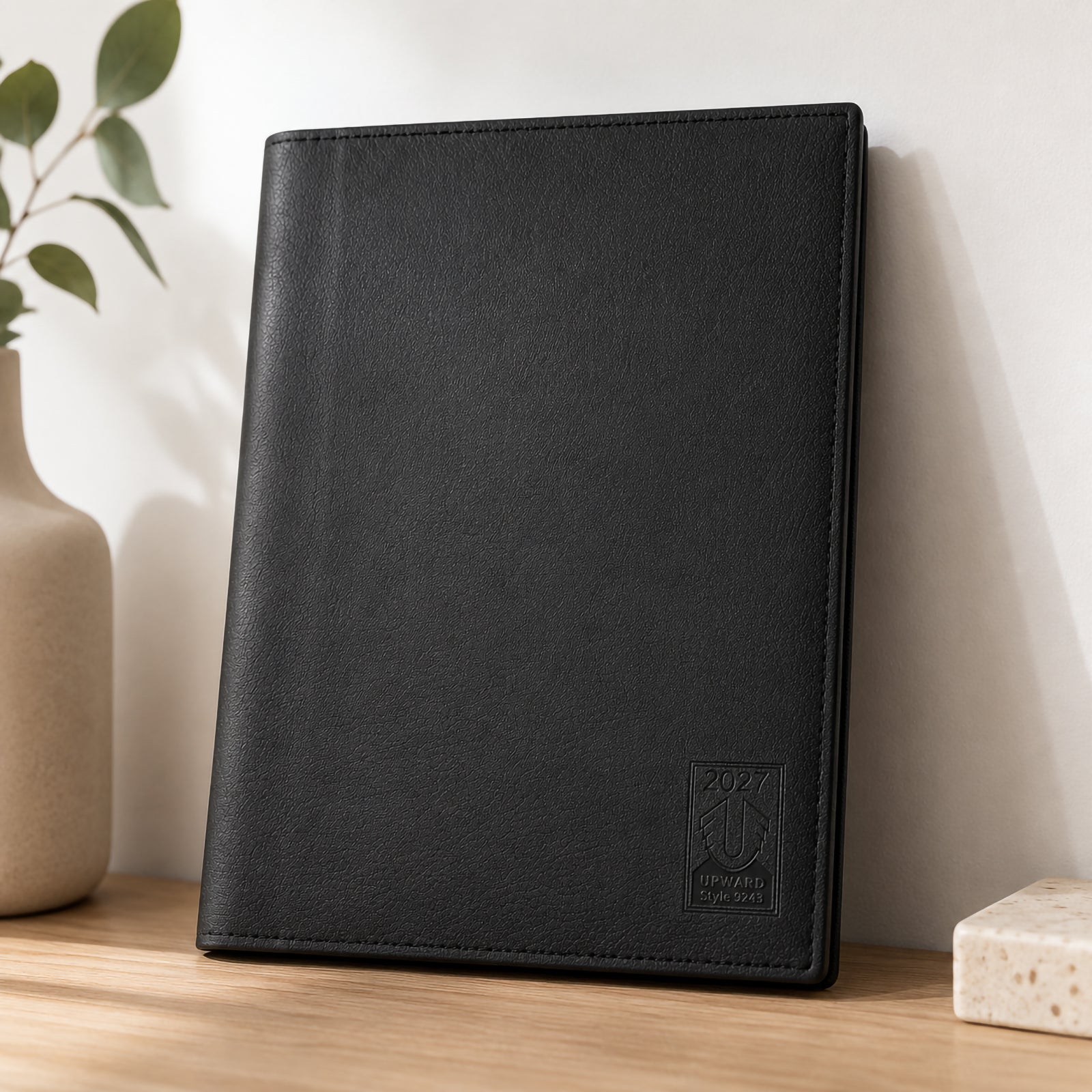 The diary stands upright on a clean office desk, showcasing its elegant textured black PU cover with subtle embossed logo. Minimalist décor creates a sophisticated business setting, emphasising durability, professionalism and timeless design.