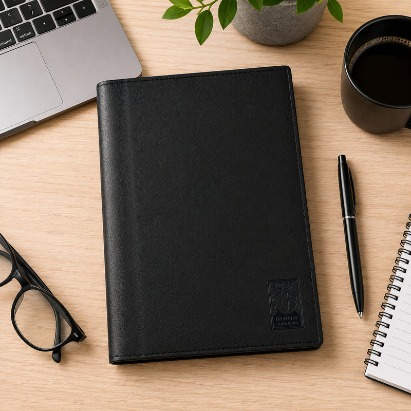 Overhead workspace view of the closed Upward Nobel 2027 A4 Diary resting beside a laptop, glasses, notebook, coffee and pen. The large black PU cover and executive styling demonstrate its suitability for professional planning, office organisation and daily desk use.