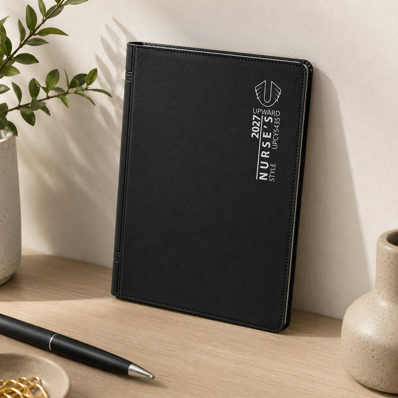 The diary standing upright on a shelf with office accessories and greenery nearby. The durable black PVC cover offers everyday protection while presenting a clean, professional look ideal for workplace organisation.