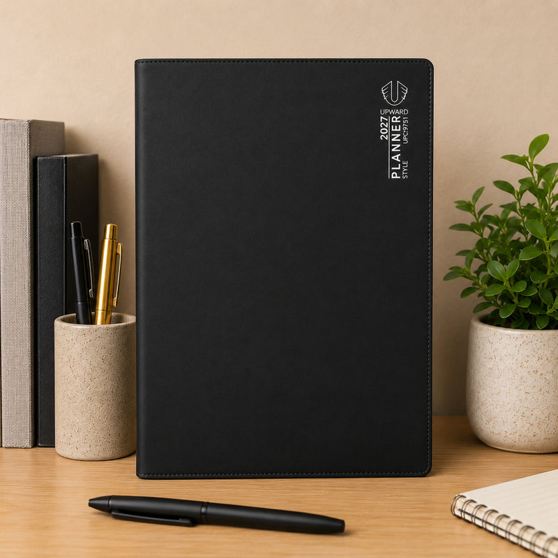 Front standing display of the Upward Planner 2027 A4 Month To View Diary in black. Set amongst books, writing instruments and a decorative plant, the planner highlights its professional design and practical format for managing deadlines, meetings and long term plans.