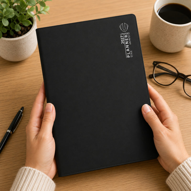 Hands hold the Upward Planner 2027 A4 Month To View Diary featuring a sleek black cover on a timber desk. Surrounded by a coffee mug, pen, eyeglasses and indoor plant, the planner presents a professional planning solution for organising appointments, monthly schedules and important dates throughout 2027.