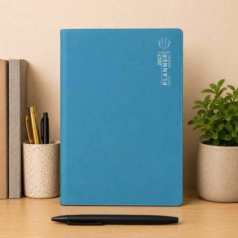 Front standing display of the Upward Planner 2027 A4 Month To View Diary in blue. Surrounded by books, writing instruments and a decorative plant, the planner presents a professional and organised look suitable for office desks, home workspaces and study areas.