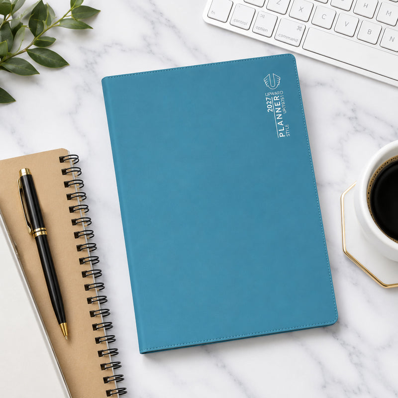 Overhead workspace view showing the Upward Planner 2027 A4 diary with a blue cover placed on a marble desk alongside a keyboard, notebook, coffee cup and pen. The clean design and spacious format make it ideal for business planning, study schedules and personal organisation.