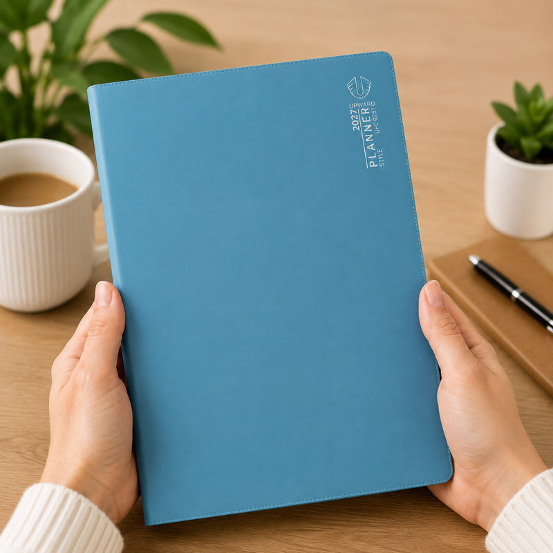 A person holds the Upward Planner 2027 A4 Month To View Diary featuring a sophisticated blue cover. Positioned on a timber desk beside a coffee mug, notebook, pen and indoor plants, the planner highlights its professional appearance and suitability for organising monthly schedules, appointments and important dates throughout 2027.