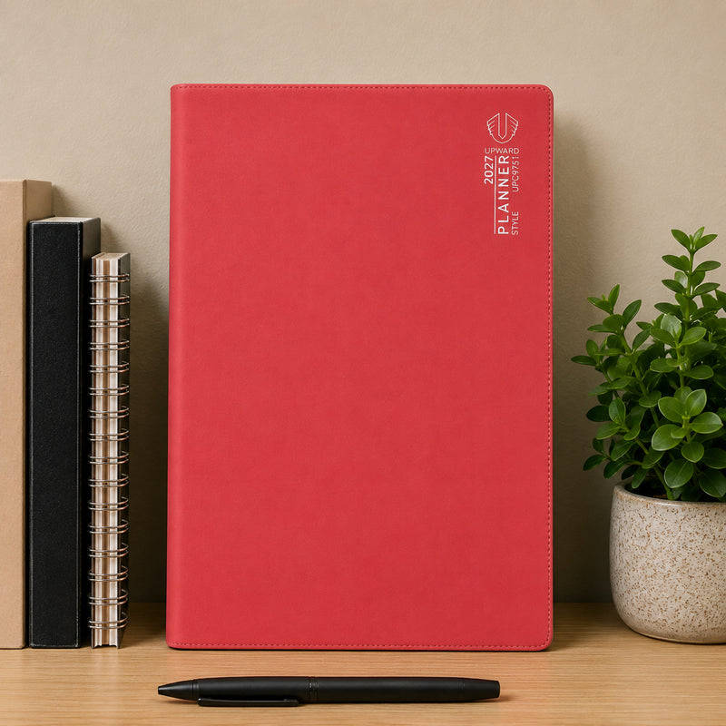 Front standing view of the Upward Planner 2027 A4 Month To View Diary in red. Displayed on a desk alongside notebooks, a pen and decorative plant, highlighting its clean design, durable cover and practical format for tracking schedules, deadlines and events.