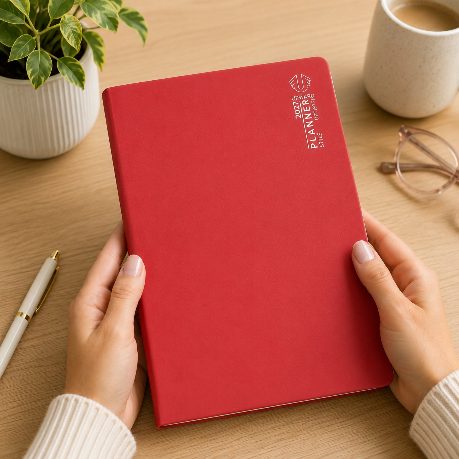 Hands hold the Upward Planner 2027 A4 Month To View Diary featuring a vibrant red cover and clean professional design. Positioned on a timber desk beside a coffee cup, pen, glasses and indoor plant, the diary presents an organised workspace ideal for monthly planning, scheduling appointments and managing important dates throughout 2027.