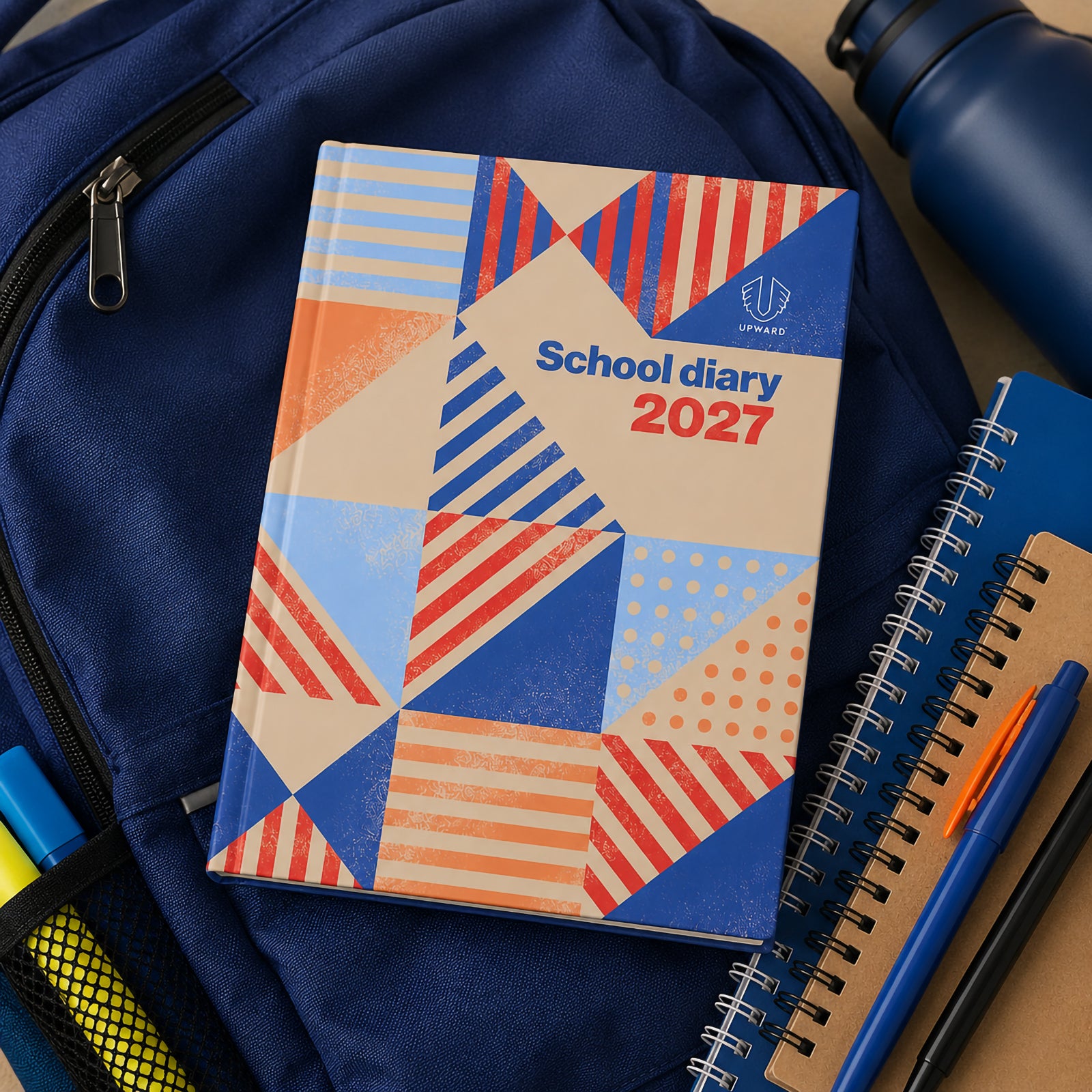 The Upward School 2027 diary displayed on a school backpack surrounded by stationery, notebooks and a drink bottle. The image demonstrates how the diary fits naturally into a student's daily routine for managing homework, assignments and schedules.