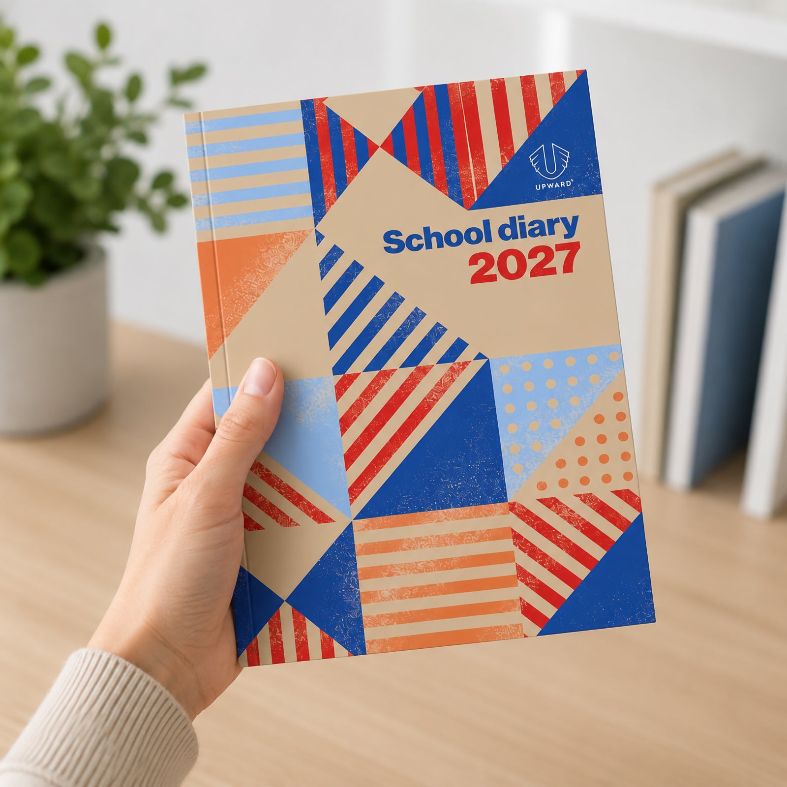 Student holding the Upward School 2027 B5 Week To View Diary featuring a colourful Geometry cover design with blue, red and orange geometric patterns. The diary is shown in a bright study environment, highlighting its portable size and student friendly appearance.