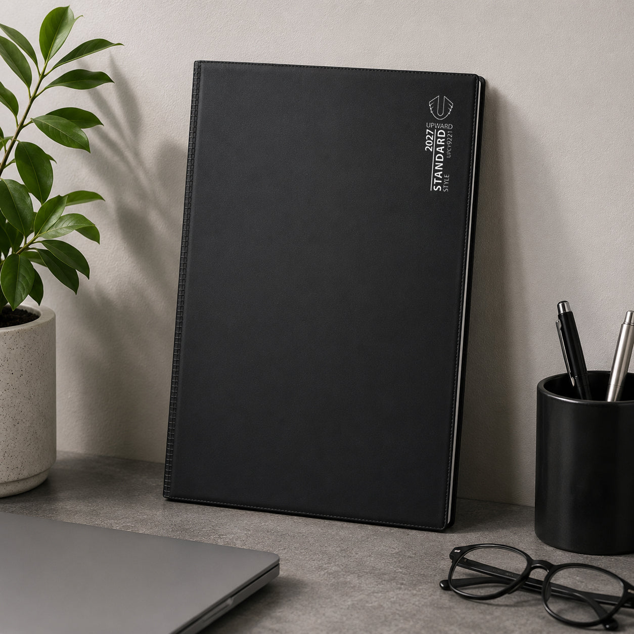 The Upward Standard 2027 A4 Day To Page Diary stands upright in a modern workspace with a laptop, stationery holder and decorative plant. Its professional appearance and spacious layout make it ideal for executive offices, study environments and home workspaces.