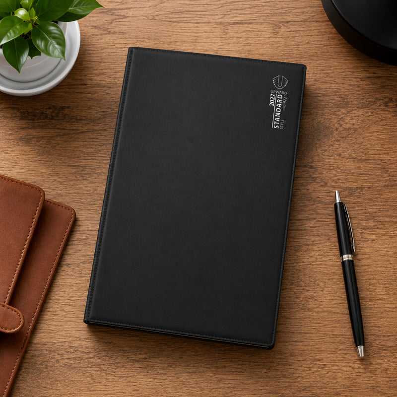 The diary is displayed on a wooden desk beside a planner wallet, pen and indoor plant, demonstrating its suitability for organised office work, business scheduling and everyday desk based planning.