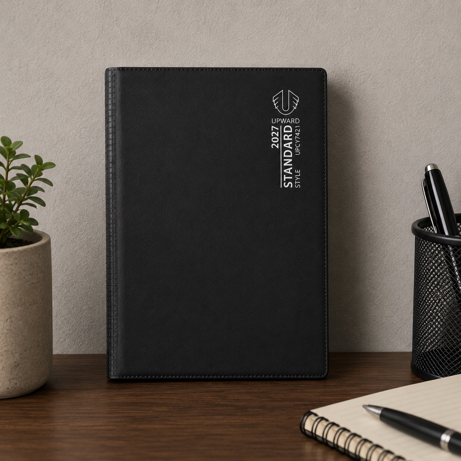 The diary stands upright on a desk next to stationery accessories and a potted plant. Its sleek black cover presents a professional and organised look, making it a practical planning tool for workplaces, home offices and study spaces.