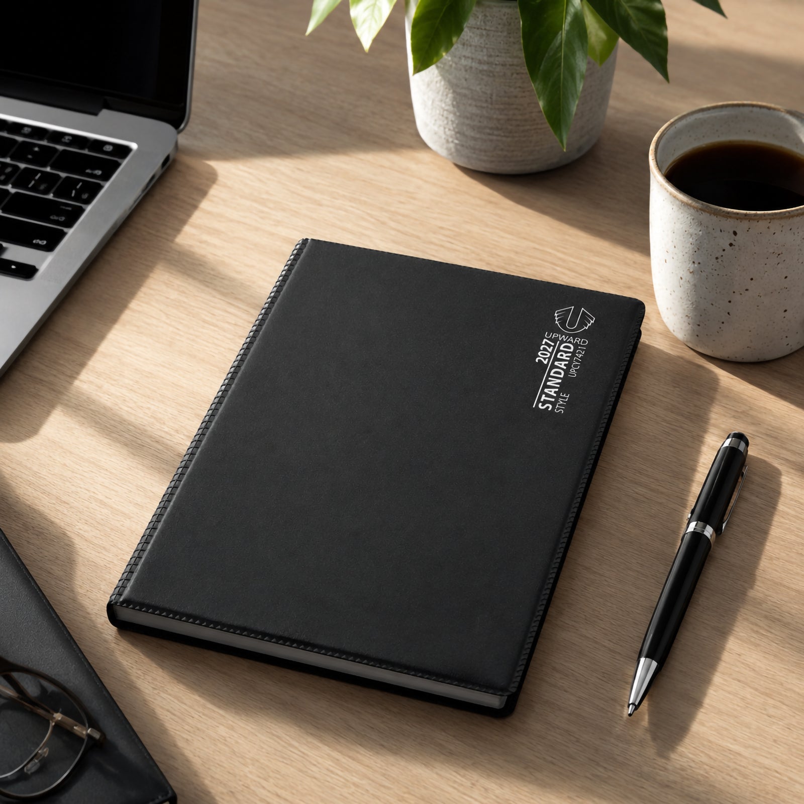 The black Upward Standard 2027 A5 Diary is displayed on a desk beside a laptop, coffee cup and pen. The professional appearance and durable cover highlight its suitability for office environments, business planning and everyday organisation.
