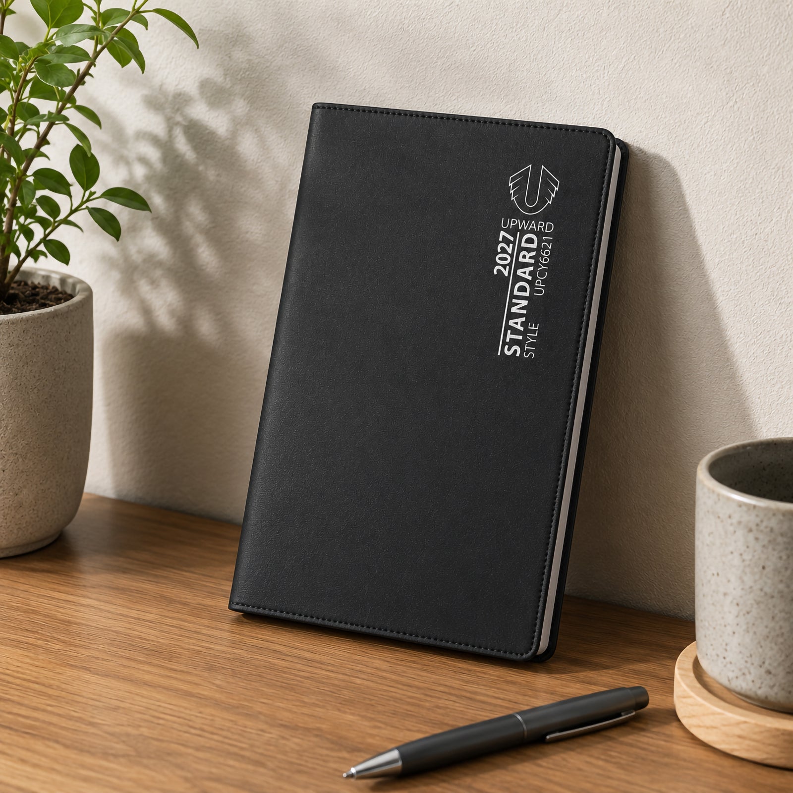 The diary stands upright on a desk next to office accessories and greenery. Its durable black cover and compact A6/7 format present a professional planning solution for students, professionals and anyone who prefers handwritten organisation.